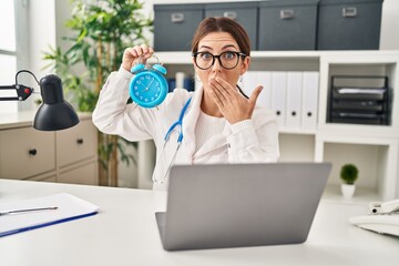 Sticker - Young brunette doctor woman holding alarm clock covering mouth with hand, shocked and afraid for mistake. surprised expression