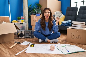 Wall Mural - Young brunette woman sitting on the floor at new home holding savings celebrating crazy and amazed for success with open eyes screaming excited.