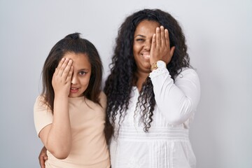 Sticker - Mother and young daughter standing over white background covering one eye with hand, confident smile on face and surprise emotion.