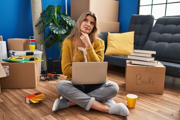 Poster - Young woman sitting on the floor at new home using laptop with hand on chin thinking about question, pensive expression. smiling with thoughtful face. doubt concept.