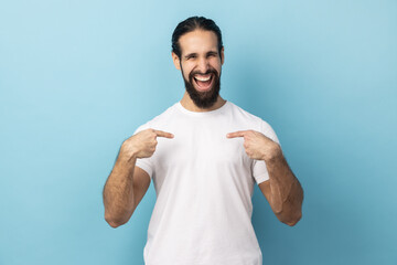 Portrait of delighted man with beard wearing white T-shirt pointing fingers on himself and looking at camera with toothy smile, extremely satisfied. Indoor studio shot isolated on blue background.