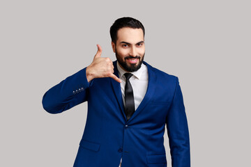 Positive bearded man standing with telephone hand gesture and smiling to camera, flirting offering to contact by phone, wearing official style suit. Indoor studio shot isolated on gray background.