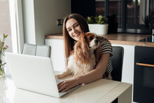 Glad, Cheerful, Smiling Young Brown Haired Woman Sit In Kitchen With Dog Cavalier King Charles Spaniel, Work On Laptop