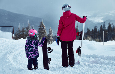 Back view of mother and daughter with snowboards looking mountains landscape together or new way to ride on ski resort at sunny winter day, travel vacation, landscape mountains background.