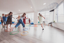 Small Nursery School Children With Female Teacher On Floor Indoors In Classroom, Doing Exercise. Jumping Over Hula Hoop Circles Track On The Floor.
