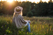 Relaxation outdoors at sunset. Boho woman with hat and poncho sitting in meadow. Enjoyment of indian summer in nature