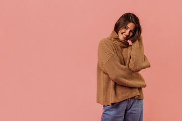 Pretty young caucasian woman wears brown sweater and jeans on pink background with place for text. Dark-haired beauty smiles looking at camera. Mood, lifestyle, concept .