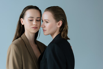 Wall Mural - young lesbian couple in beige and black blazers standing isolated on grey.