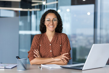 Wall Mural - Portrait of successful young businesswoman, Hispanic woman working inside modern office building smiling and looking at camera sitting at desk with laptop, female worker with curly hair and glasses