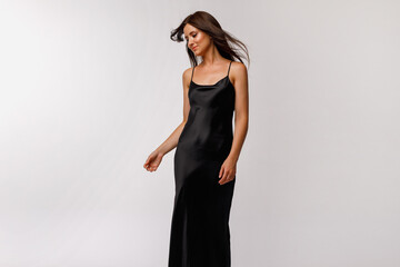 A brunette girl with long hair is standing in a black slip dress on a light background