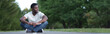 © lenblr - Young man wearing white t-shirt and jeans looks around sitting cross legged on asphalt against green trees. African American guy enjoys resting in city park, copyspace