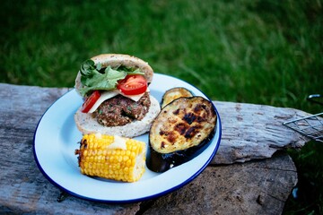 Wall Mural - Fresh tasty homemade beef burger, eggplant barbecue and corn cobs on plate in park on wooden bench