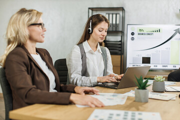 Wall Mural - Side view of focused caucasian woman talking using headset while her female colleagues listening speech of african american business woman. Company managers working at office room. Deadlines concept.