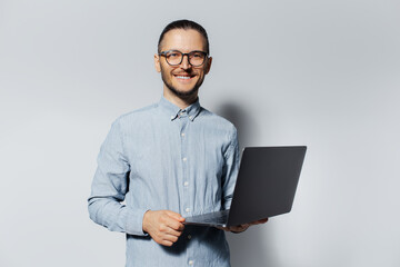 Wall Mural - Studio portrait of young smiling man holding a laptop in hands, wearing blue shirt and eyeglasses on white background.