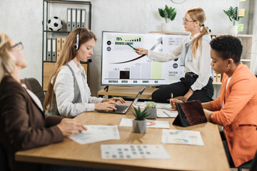 Group of multi ethnic female partners sitting at desk and listening speech of caucasian woman near monitor with various graphs and charts. Business meeting at office.