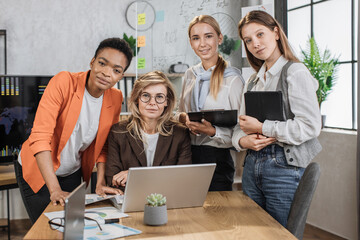 Wall Mural - Beautiful aged senior female boss sitting at the table in office and listening to her diverse colleagues, standing around and discussing results of joint work or strategy.