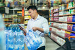 © Liubomir - Wholesale purchase. A serious young Asian man buys a pallet of mineral water in a supermarket. Holds the phone in his hand, looks at the phone, uses the self-service.