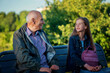 Granddad and granddaughter sitting on wooden bench at city park against nature background and talking