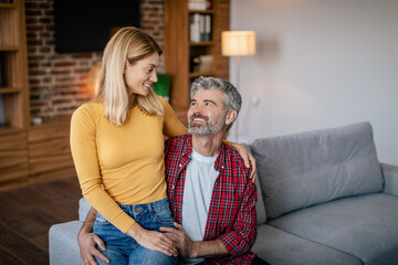 Wall Mural - Calm smiling mature european wife sits on husband knees, rest, enjoy free time on sofa in cozy living room interior