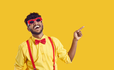 Funny extravagant Indian guy on orange background is advertising new promotion or offering sale. Man in yellow shirt, red suspenders, bow tie and funny glasses laughs while pointing his finger to side