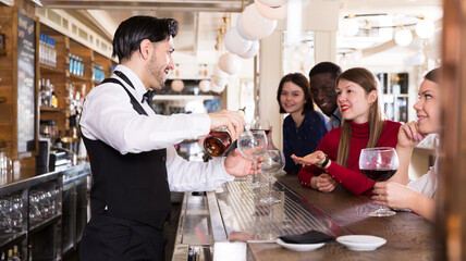 Wall Mural - Smiling bartender pouring beverages and talking to cheerful visitors in bar.