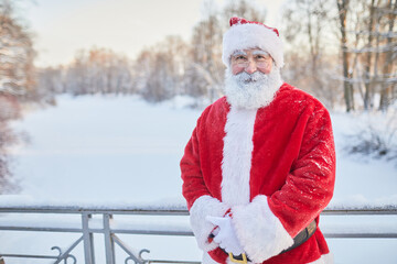 Wall Mural - Waist up portrait of traditional Santa Claus looking at camera and smiling in winter park, copy space