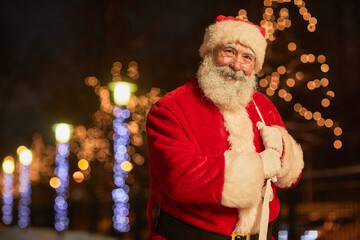 Wall Mural - Waist up portrait of traditional Santa Claus holding sack with presents and smiling at camera in night city with lights, copy space