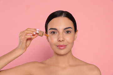 Poster - Young woman applying essential oil onto face against pink background