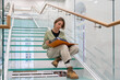 Focused mature female reader sits on modern glass stairs step enjoying interesting book in contemporary public library. Curious woman in casual outfit reads about world history getting new knowledge