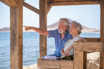 Cheerful caucasian senior couple sitting in the shade of the gazebo in front of the sea looking away - elderly pensioners enjoying sea vacation in a windy sunny day