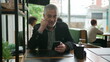 © Marco - Older man using cellphone inside coffee shop watching content online on phone. A middle aged person looking at smartphone screen