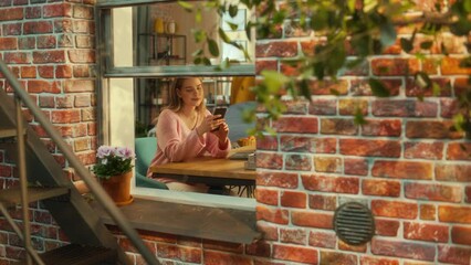 Poster - Positive, Smiling Beautiful Female Using a Smartphone while Sitting Behind a Table in Cozy Living Room. Girl Checking Online Social Media. Cinematic Shot from Outside the Building.