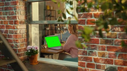 Fototapete - Female Working on a Laptop Computer with Green Screen Mock Up Display while Sitting Behind a Table in Stylish Living Room. Business Manager Working Remotely from Home. View from Outside the Window.