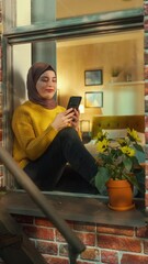 Poster - Vertical Screen: Muslim Girl Wearing a Hijab, Using a Smartphone while Sitting on a Window Sill in Brooklyn Style Brownstone House. Young Female Browsing Internet. Camera Angle from Outside the House.