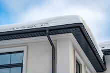 Metal Downpipe System, Guttering System, External Downpipes And Drainage Pipes Under Snow. Corner Of House With Roof Made Of Gray Metal Tiles And Gutter Covered With Thick Layer Of Snow In Winter.