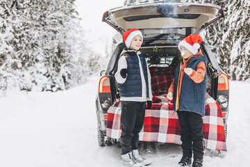 Wall Mural - Two friends teenagers boys in red santa hats with with sparkles Bengal fire standing near trunk of car decorated for Christmas and New Year in snowy winter forest. Road trip and local travel.