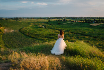 Bride holding her wedding dress and running in a field on the road with sunlight. Woman in the mountains top at sunset. Girl walking on meadow in green grass. Back view.