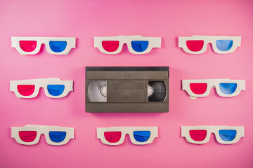 Wall Mural - Old white paper 3d glasses with blue red lenses and black videotape on pink background. Retro video cassette with magnetic tape and stereoscopic 3D cardboard glasses. Vintage background.