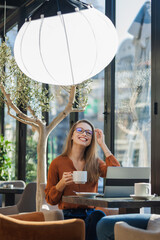 Young female model posing as businesswoman in a cafe bar with lantern softbox light equipment. Freelancer girl working on laptop and having tea at a window table..