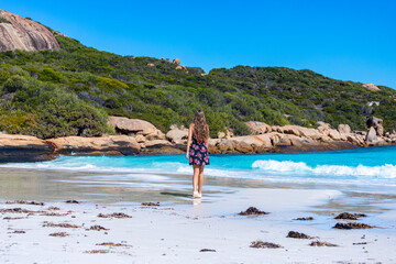 a long-haired girl in a black dress with roses walks along a paradise beach with white sand and turquoise water and orange rocks, cape le grand national park near esperance, western australia