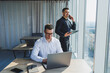 © DSMT - Two male focused colleagues discussing business project looking at laptop screen during meeting in modern meeting room with glass walls