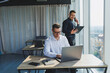 © DSMT - Two male focused colleagues discussing business project looking at laptop screen during meeting in modern meeting room with glass walls