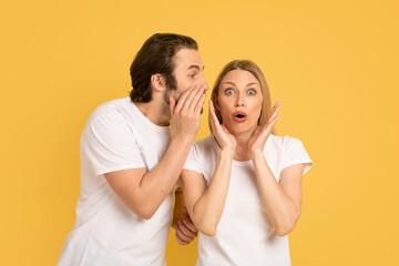 Cheerful young caucasian guy whispering into ear of shocked woman in white t-shirt