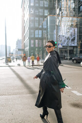 Wall Mural - Girl on the street in a black coat. Stylish girl in a black raincoat on the background of a high glass house