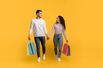 Wall Mural - Big Sales. Happy Arab Couple Walking With Shopping Bags On Yellow Background