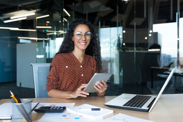 Wall Mural - Happy and successful hispanic woman working inside modern office building, business woman using tablet computer smiling and looking at camera worker doing paperwork.