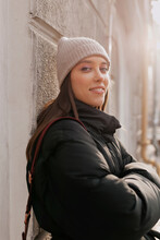 Attractive Lovable Girl With Brown Hair Wearing Knitted Cap And Black Jacket Posing And Smiling At Camera In Sunlight. Outdoor Portrait Of Elegant Young Woman Posing In Cold Day. 
