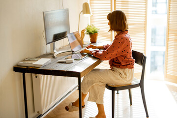 Woman works on computers while sitting by wooden table at sunny room at home. Concept of remote work from home at cozy atmosphere