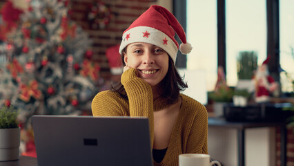 Portrait of office worker wearing santa hat during winter holiday, working on laptop at desk before celebrating christmas eve. Woman feeling festive in space decorated with xmas ornaments.