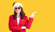 © Studio Romantic - Christmas and new year banner or announcement. Beautiful woman in santa hat and red suit pointing with index finger on copy space on orange background. New year discount advertising concept.